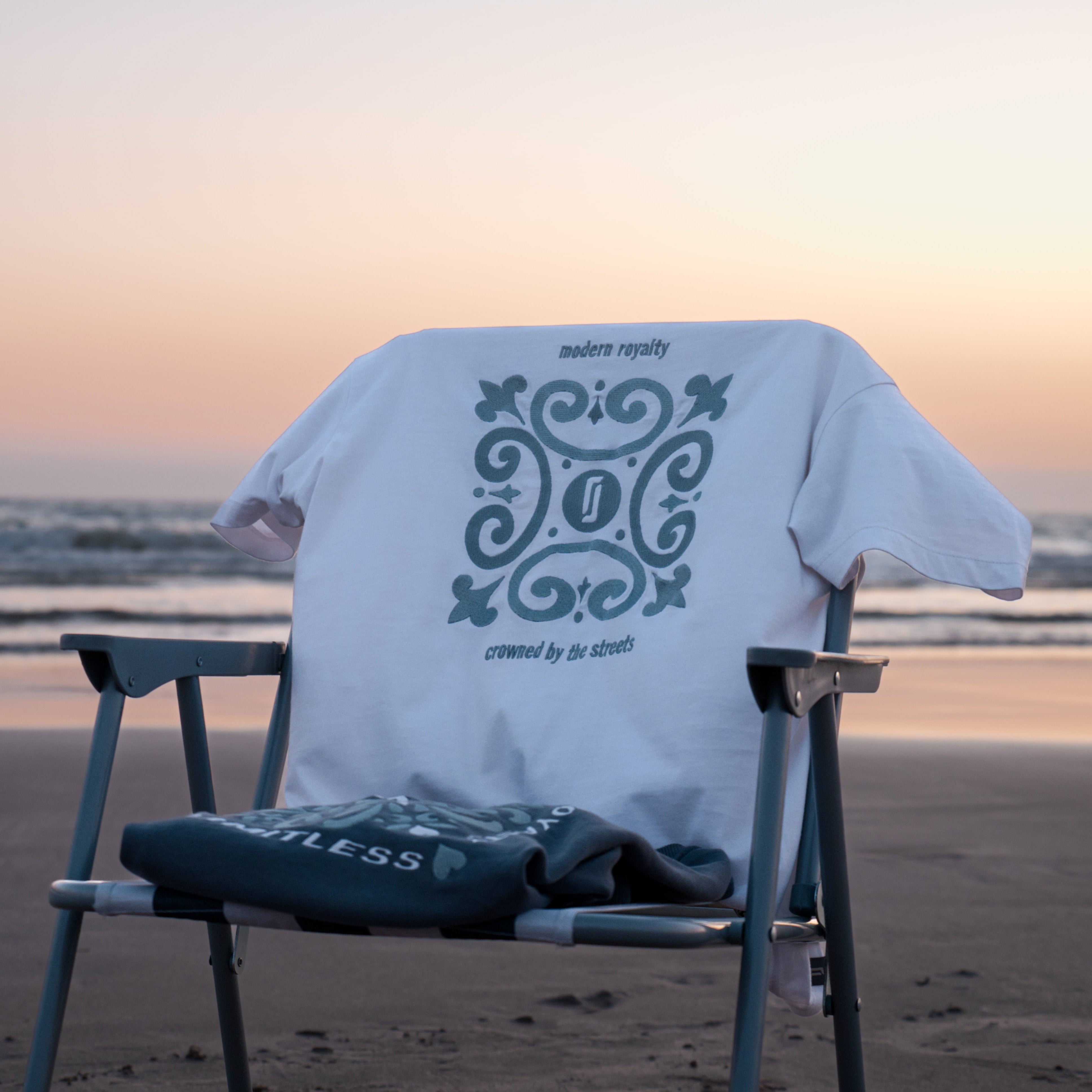 T-shirt Limitless Alvura branca com bordado Modern Royalty pendurada na cadeira e Crewneck Abissal azul profundo dobrado, fotografia premium ao pôr-do-sol na praia.