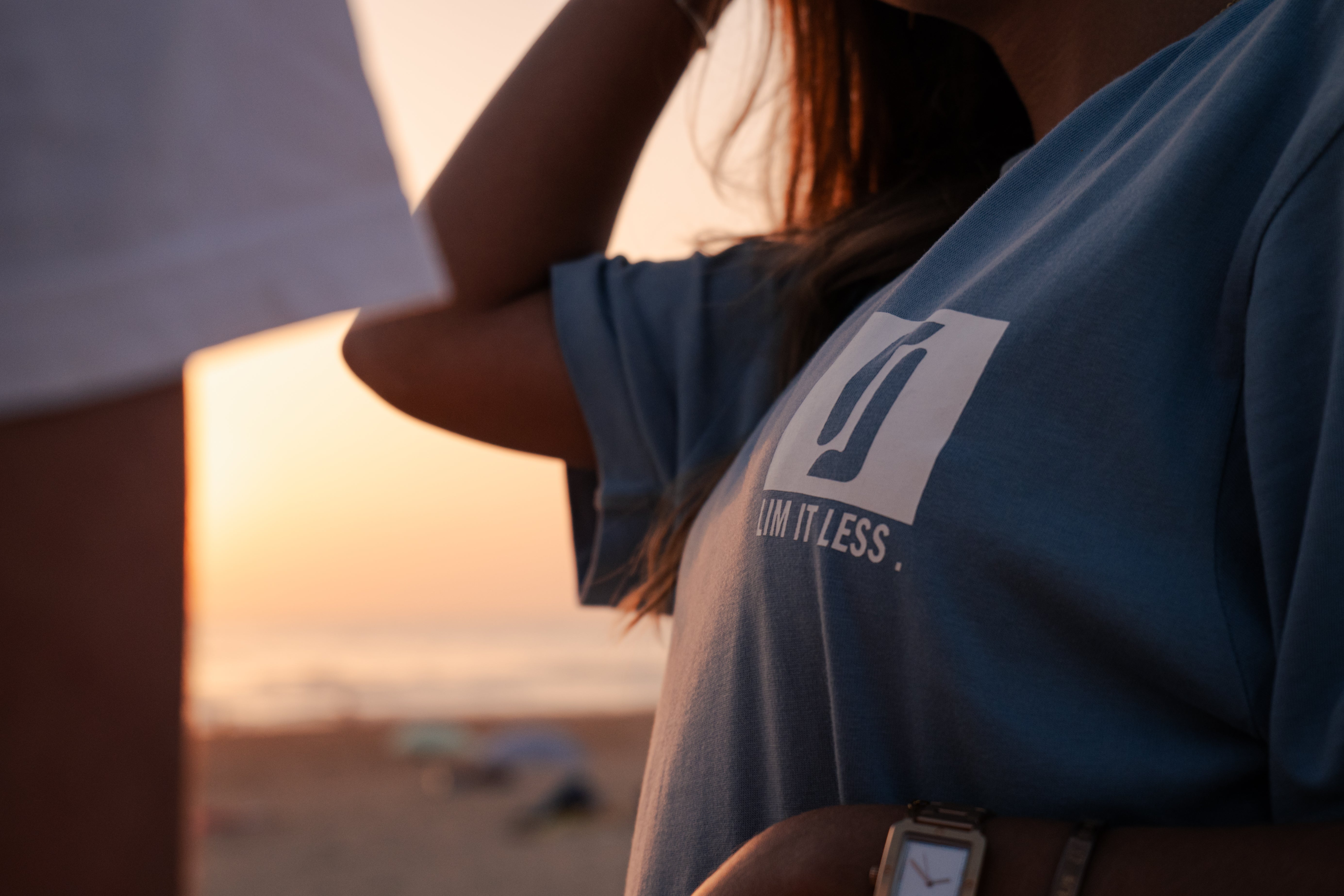 T-shirt Limitless Baía azul acinzentado em algodão premium, detalhe do logo ao pôr-do-sol na praia, com T-shirt Alvura branca em plano próximo.