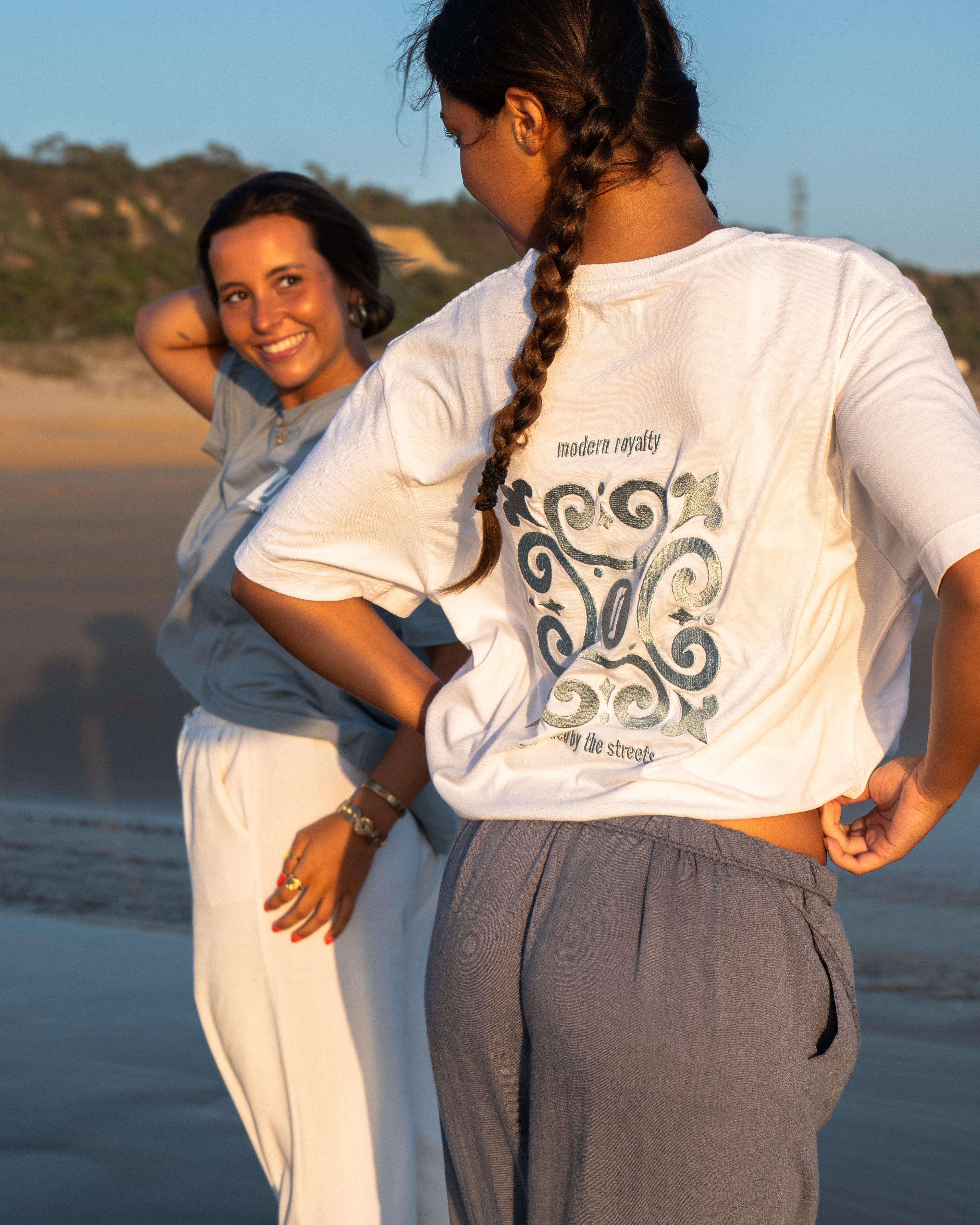 Modelos femininas com T-shirt Limitless Alvura branca com bordado Modern Royalty e T-shirt Baía azul acinzentado, fotografia aspiracional na praia ao pôr-do-sol, coleção Modern Royalty.