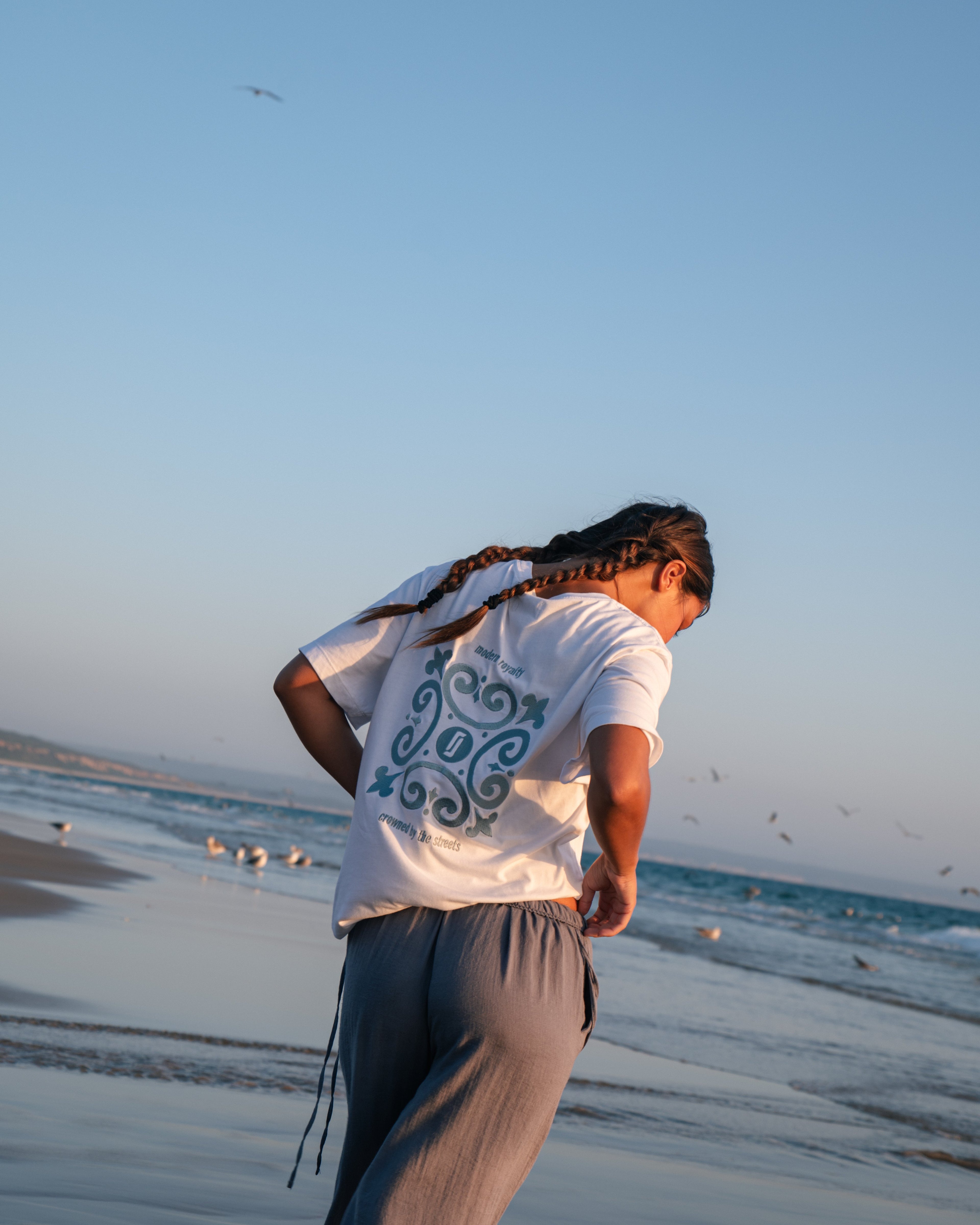 Modelo feminina com T-shirt Limitless Alvura branca em algodão premium, detalhe do bordado Modern Royalty nas costas, fotografia aspiracional na praia ao entardecer.
