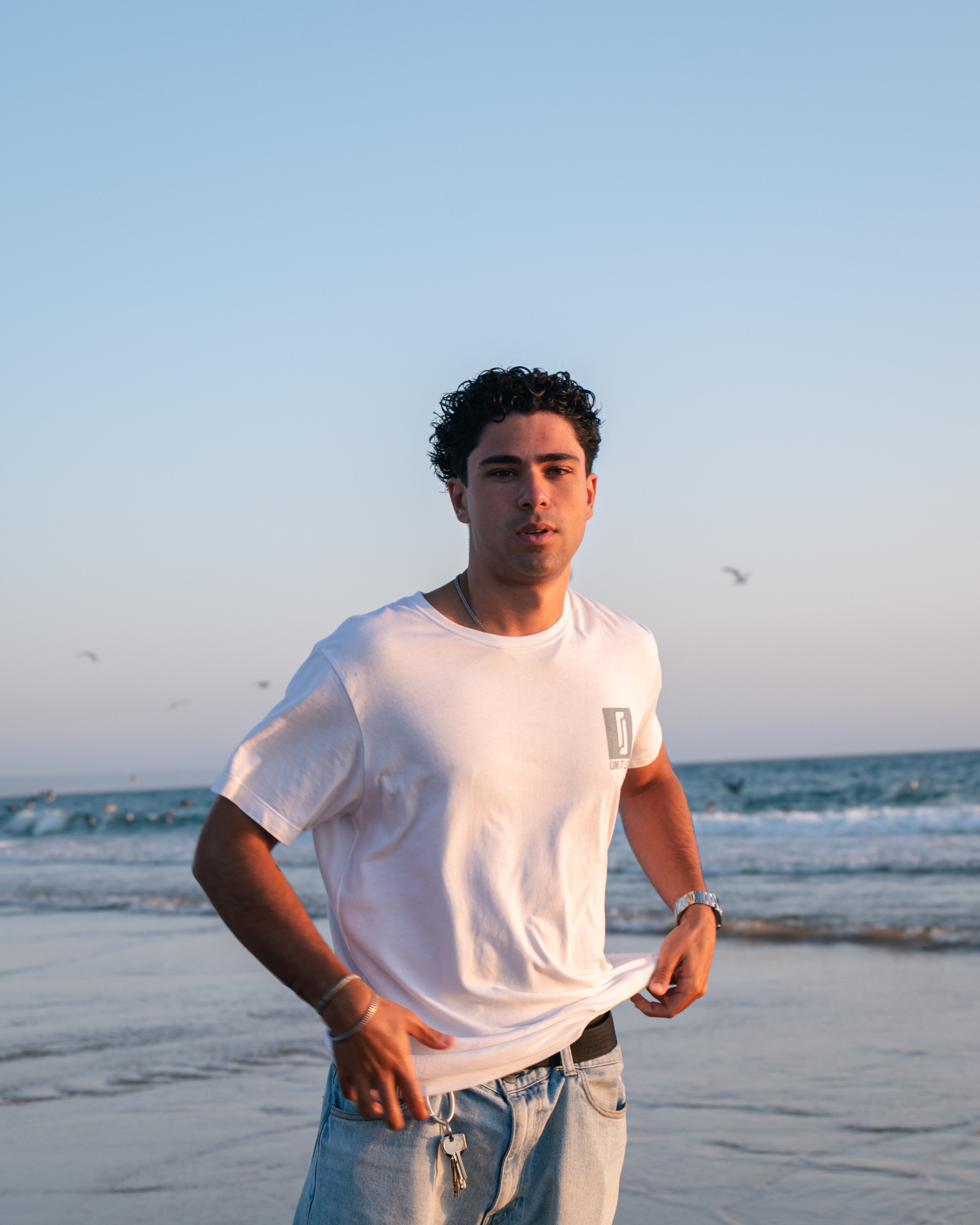 Modelo masculino com T-shirt Limitless Alvura branca em algodão premium, fotografia aspiracional na praia ao pôr-do-sol, coleção Modern Royalty.