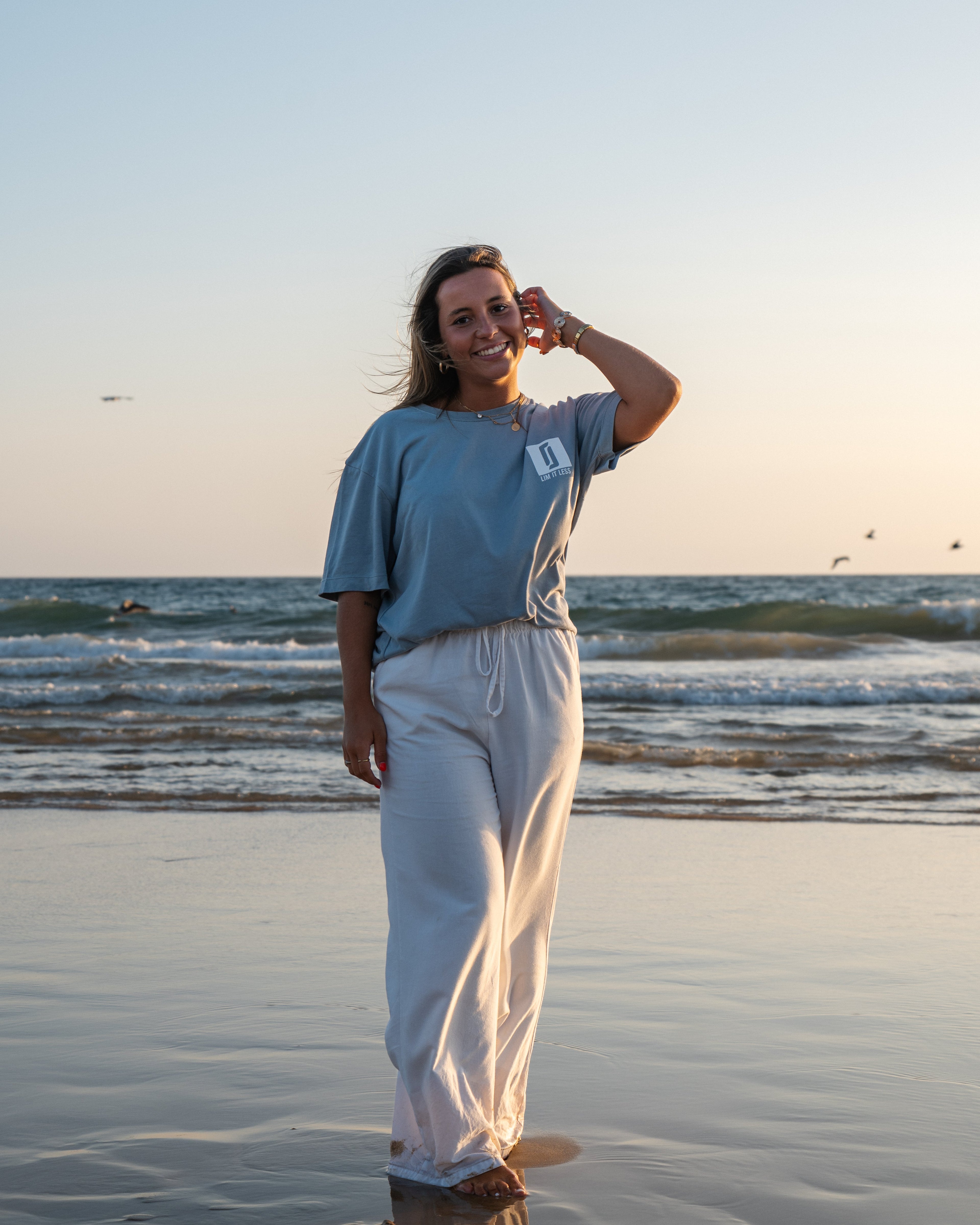 Modelo feminina sorridente com T-shirt Limitless Baía azul acinzentado em algodão premium, fotografia aspiracional na praia ao pôr-do-sol, coleção Modern Royalty.