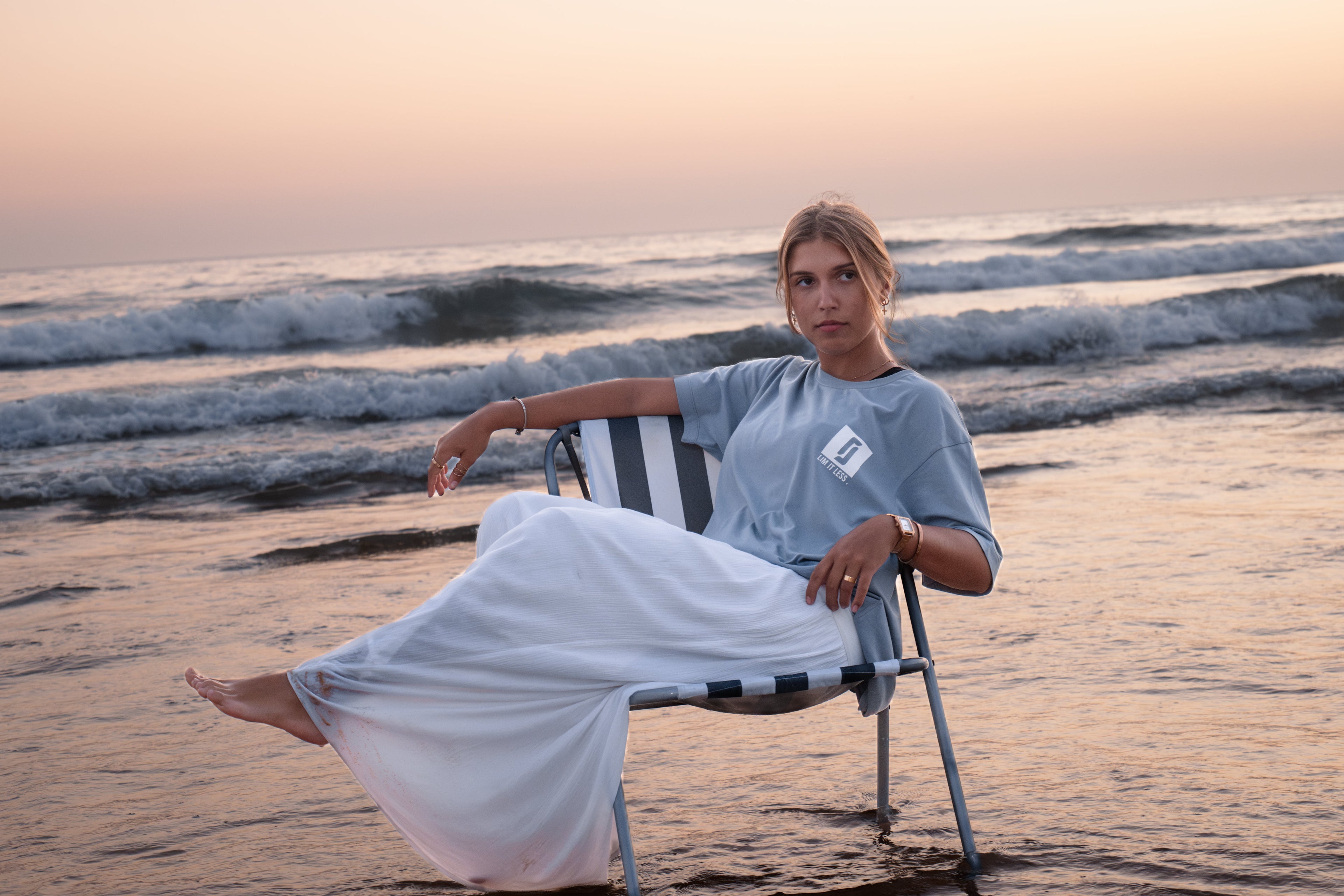 Modelo feminina com T-shirt Limitless Baía azul acinzentado sentada em cadeira na praia ao pôr-do-sol, coleção Modern Royalty com estilo premium descontraído.