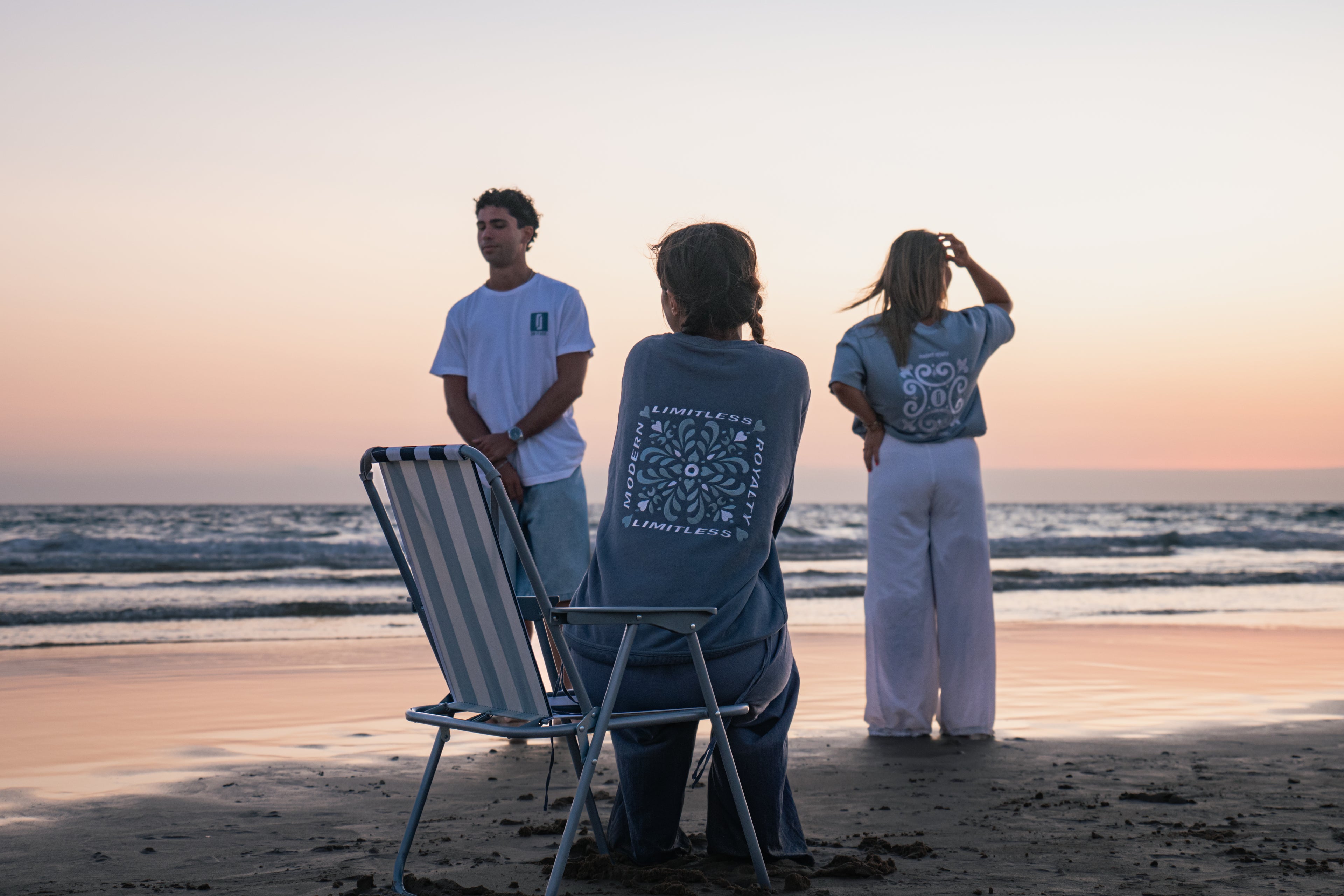 Três modelos na praia ao pôr-do-sol com peças LIMITLESS da coleção Modern Royalty: T-shirt Alvura branca, T-shirt Baía azul acinzentado e Crewneck Abissal azul profundo, fotografia aspiracional de lifestyle.