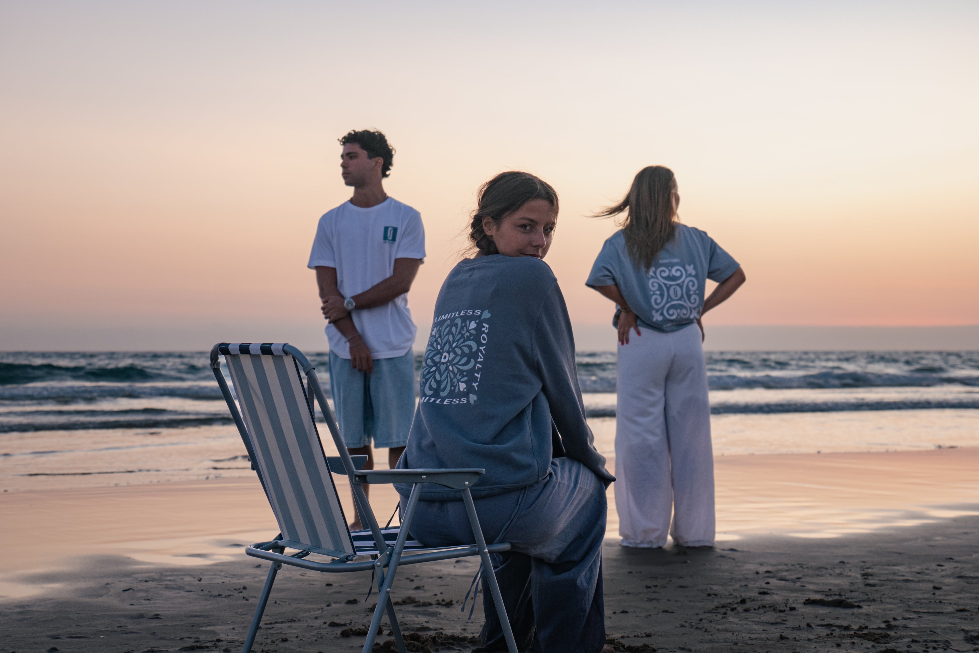 Modelos com T-shirt Limitless Alvura branca, T-shirt Baía azul acinzentado e Crewneck Abissal azul profundo, coleção Modern Royalty na praia ao pôr-do-sol.