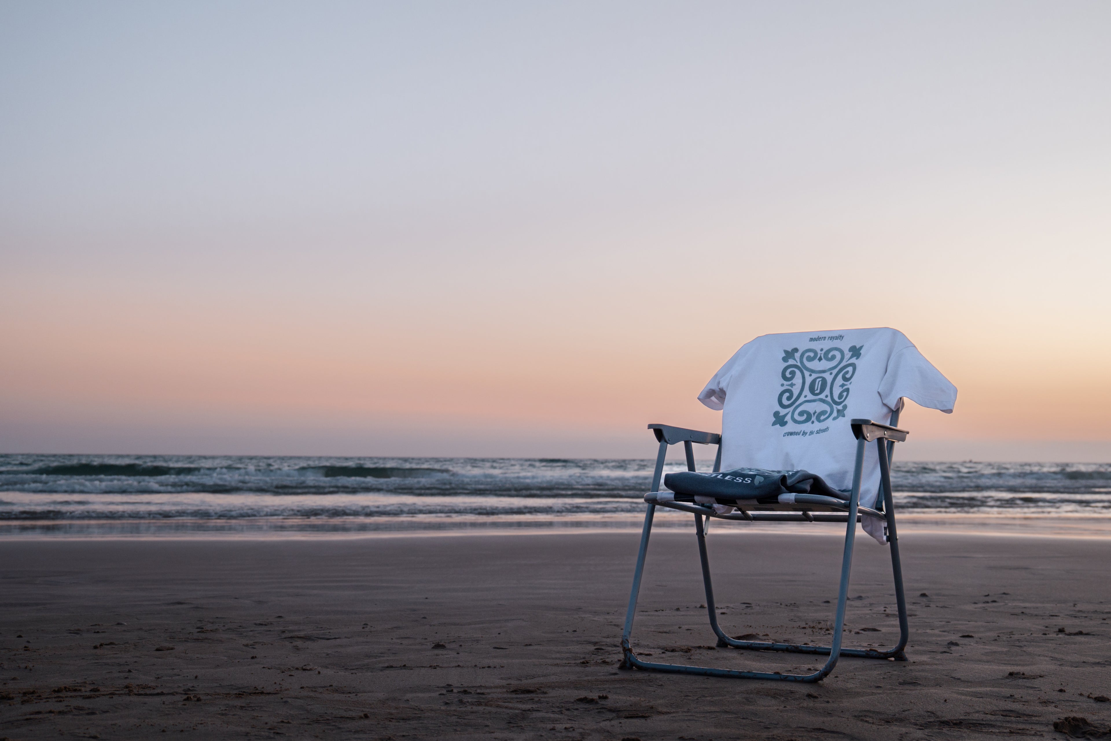 T-shirt Limitless Alvura branca com bordado Modern Royalty pendurada em cadeira e Crewneck Abissal azul profundo dobrado, fotografia premium na praia ao pôr-do-sol, coleção Modern Royalty