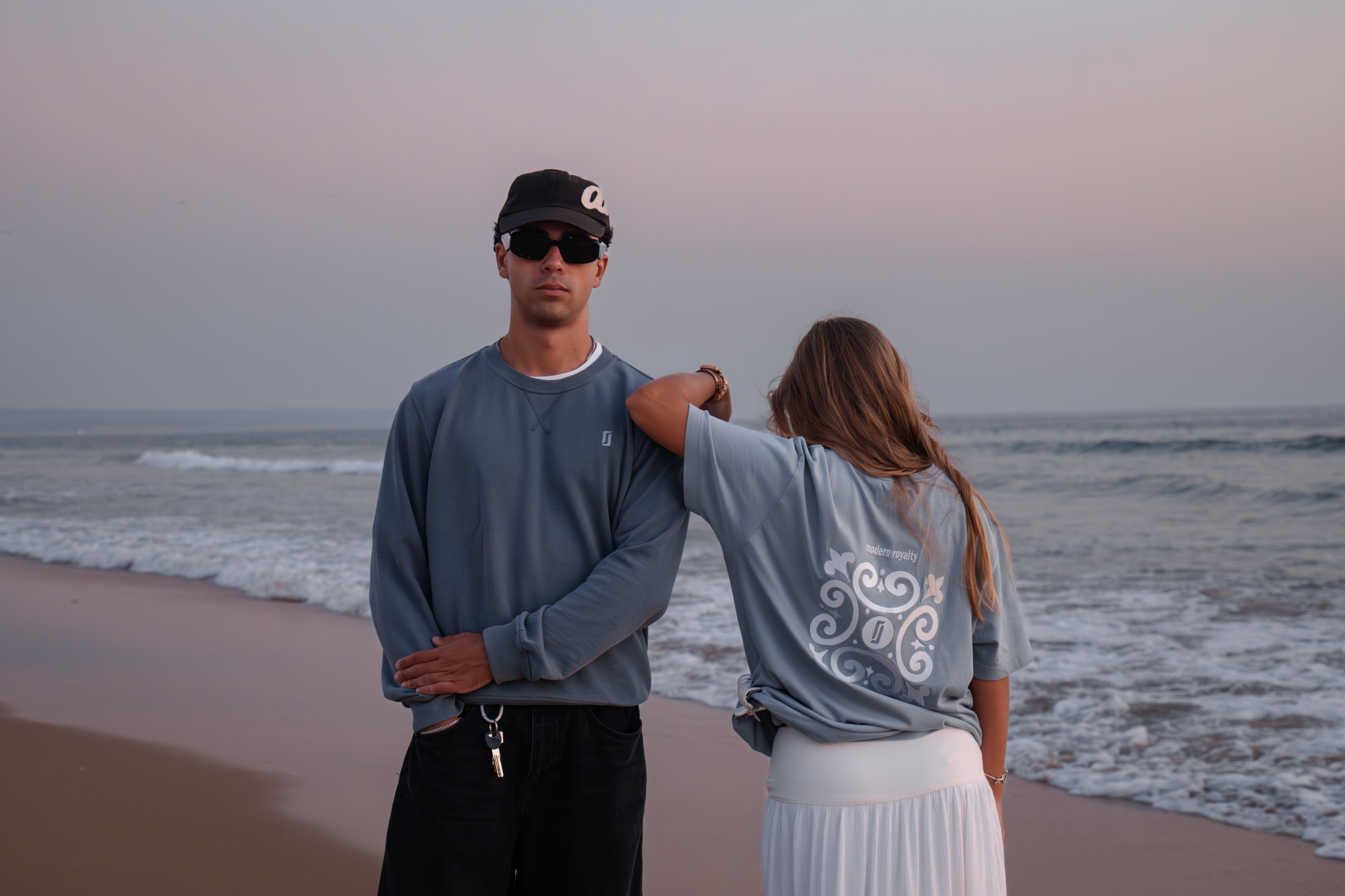 Modelo masculino com Crewneck Limitless Abissal azul profundo e modelo feminina com T-shirt Baía azul acinzentado apoiada no ombro, fotografia premium na praia ao entardecer, coleção Modern Royalty.