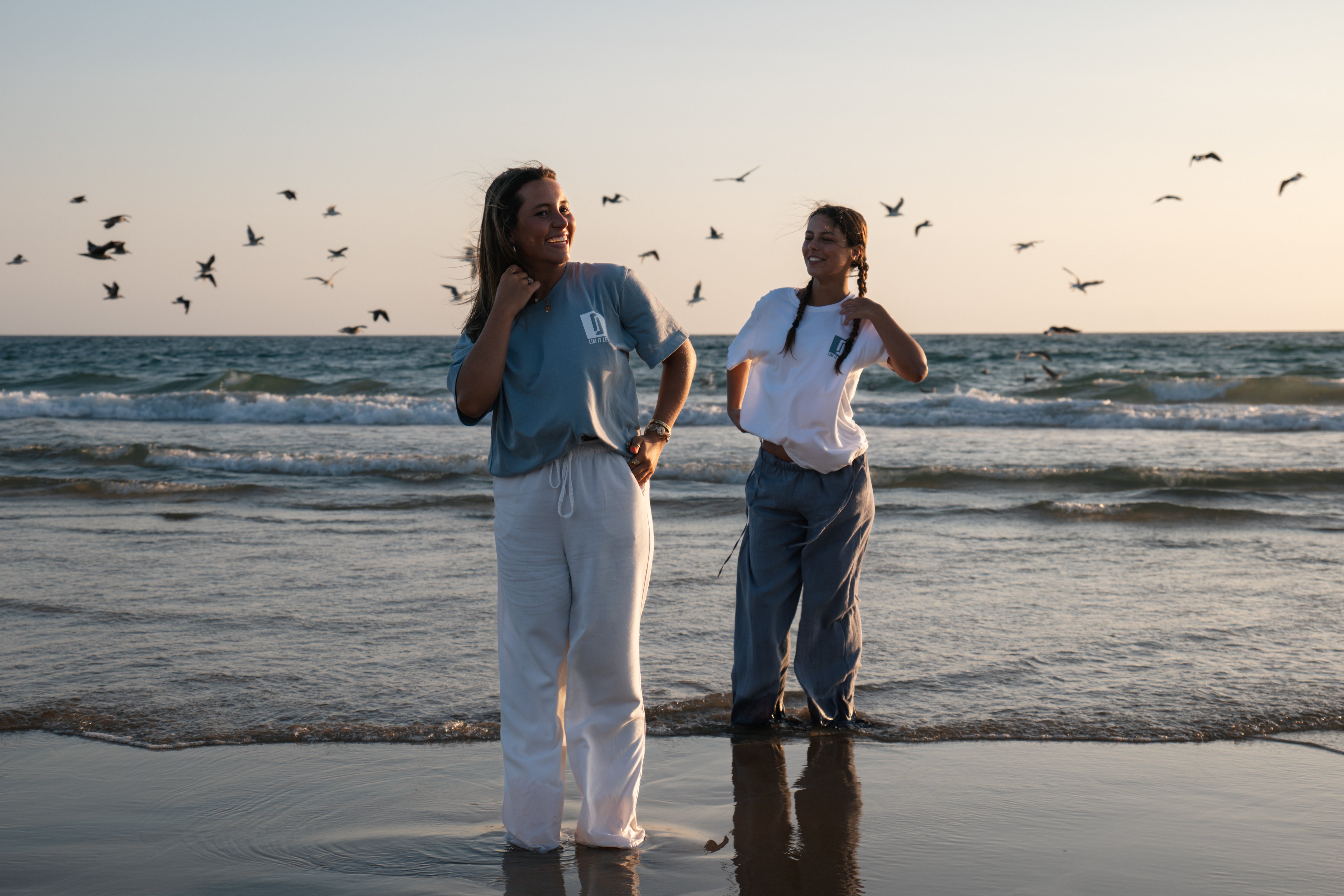Modelos femininas com T-shirt Limitless Baía azul acinzentado e T-shirt Alvura branca, fotografia aspiracional na praia ao pôr-do-sol com aves em voo, coleção Modern Royalty.