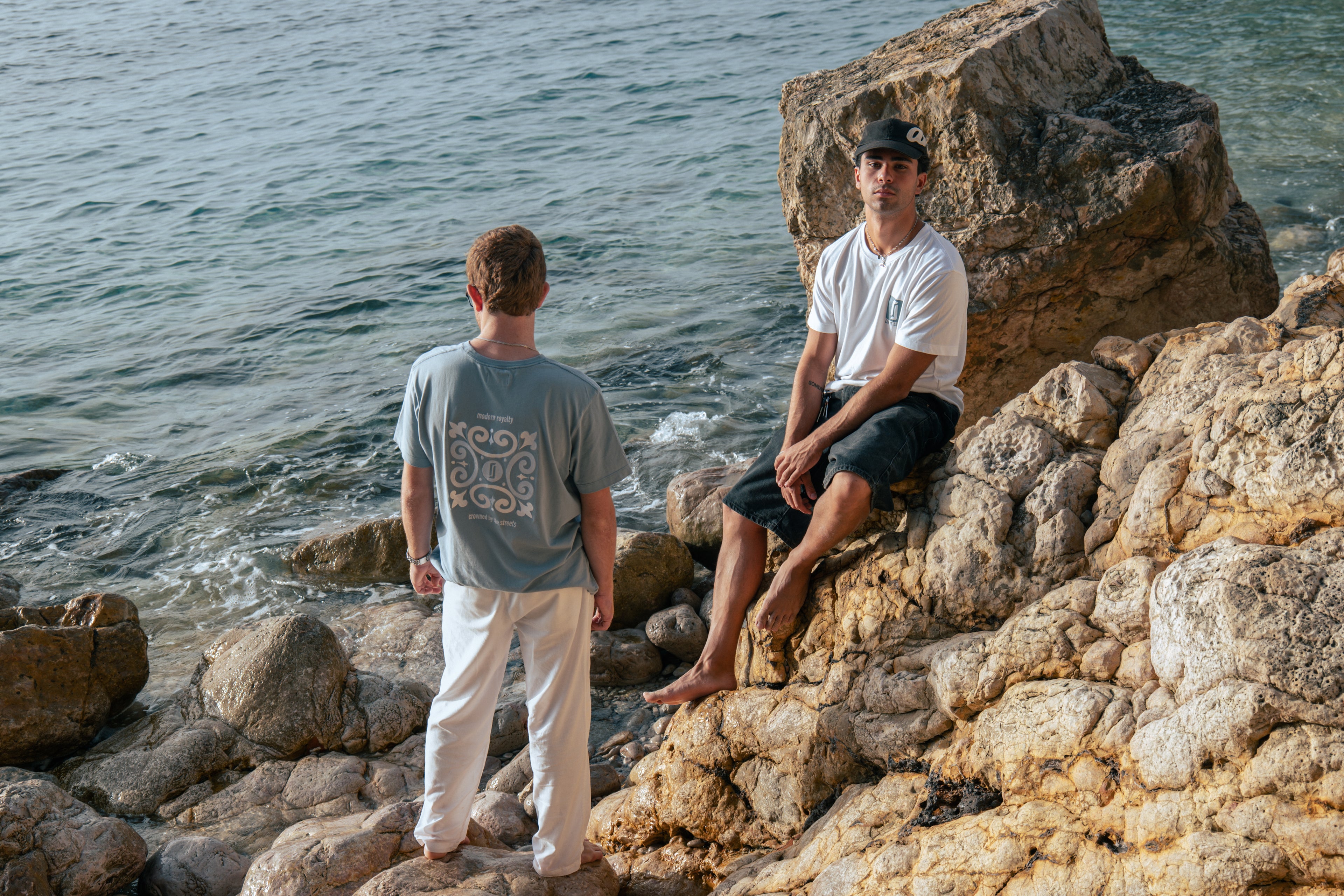 Modelos masculinos com T-shirt Limitless Baía azul acinzentado e T-shirt Alvura branca, fotografia premium em rochas junto ao mar, coleção Modern Royalty