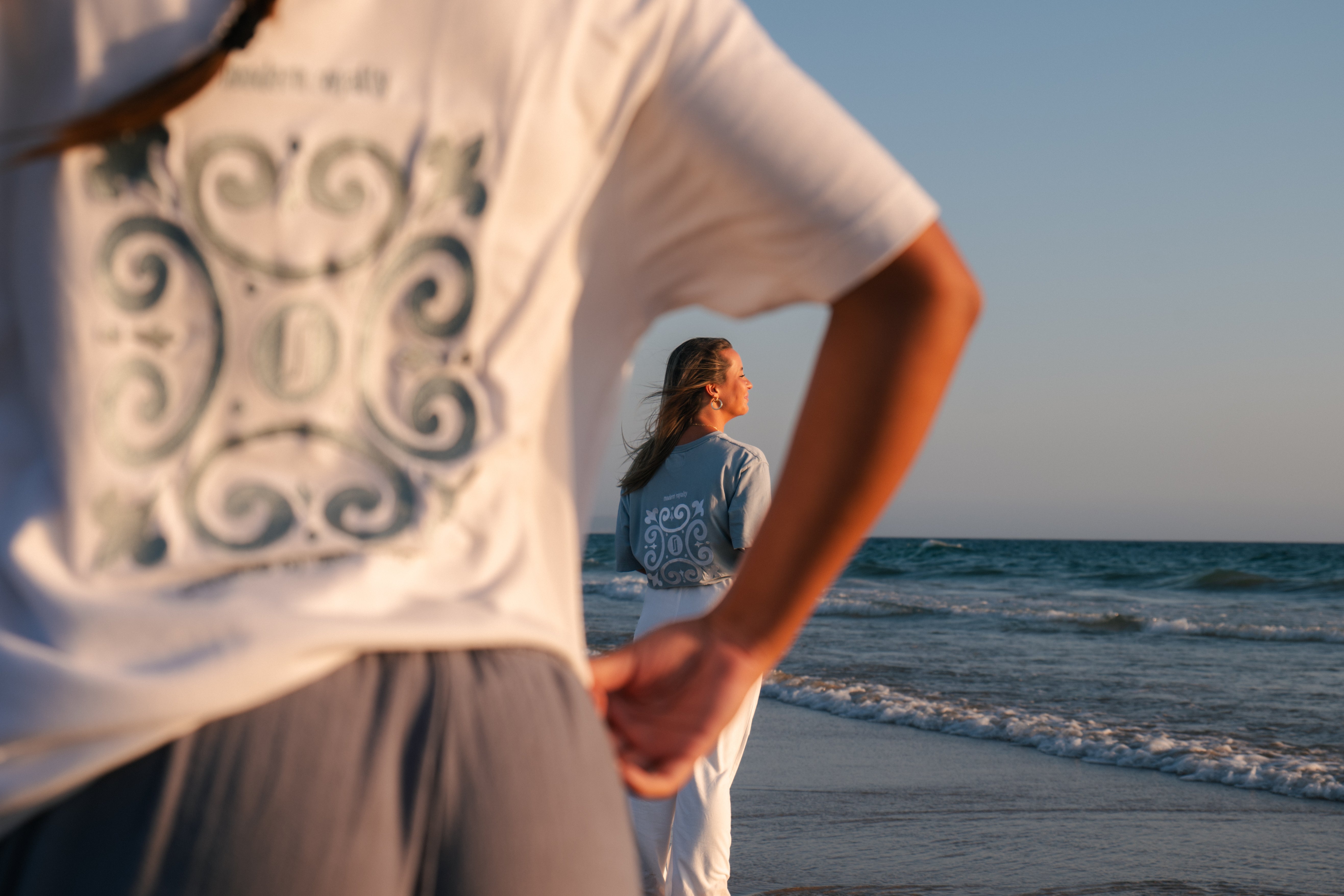 Detalhe da T-shirt Limitless Alvura branca com bordado Modern Royalty em primeiro plano e modelo feminina com T-shirt Baía azul acinzentado ao fundo na praia ao entardecer