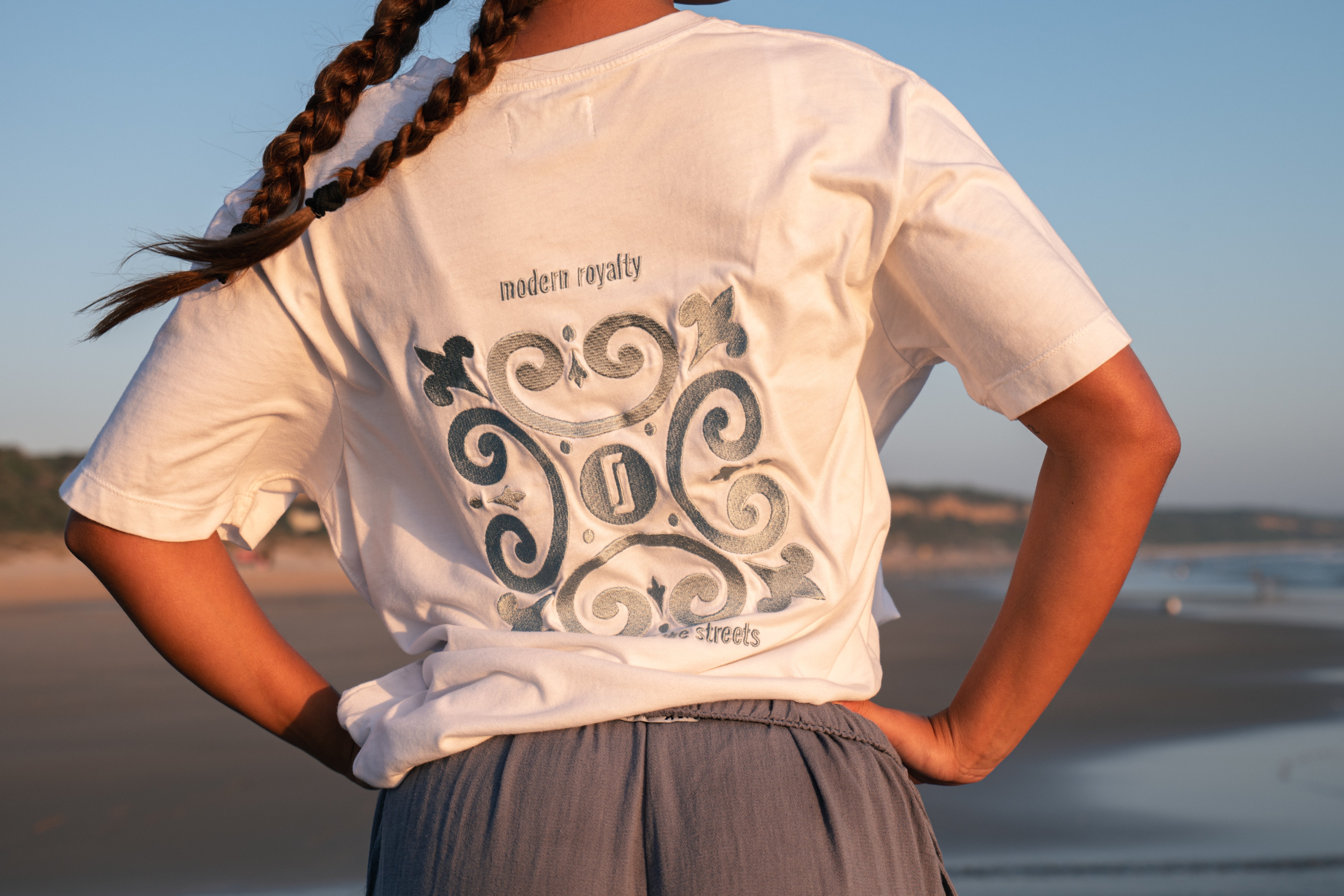 Close-up da T-shirt Limitless Alvura branca em algodão premium, detalhe do bordado Modern Royalty nas costas, fotografia minimalista na praia ao entardecer, coleção Modern Royalty