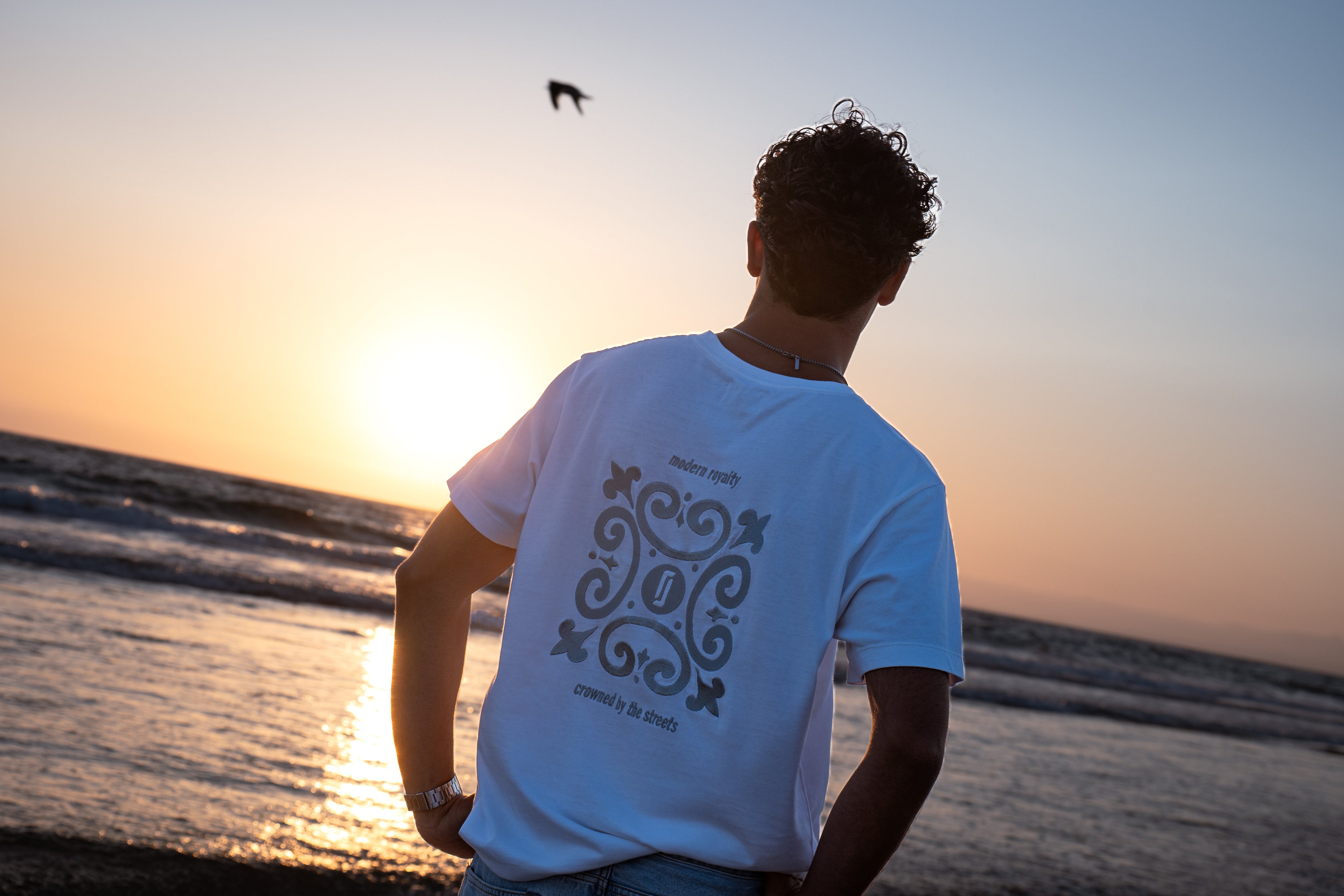 Modelo masculino com T-shirt Limitless Alvura branca e bordado Modern Royalty nas costas, fotografia aspiracional ao pôr-do-sol na praia, coleção Modern Royalty.