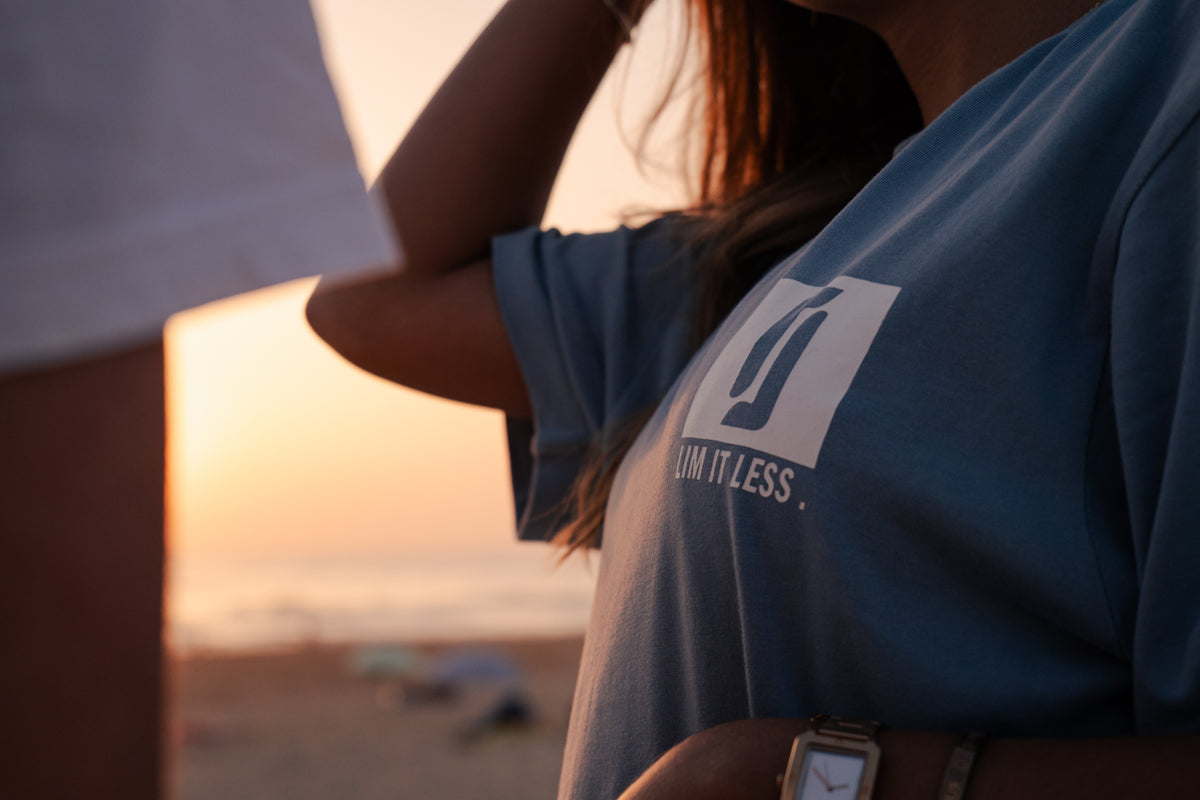 T-shirt Limitless Baía azul acinzentado em algodão premium, detalhe do logo ao pôr-do-sol na praia, com T-shirt Alvura branca em plano próximo.