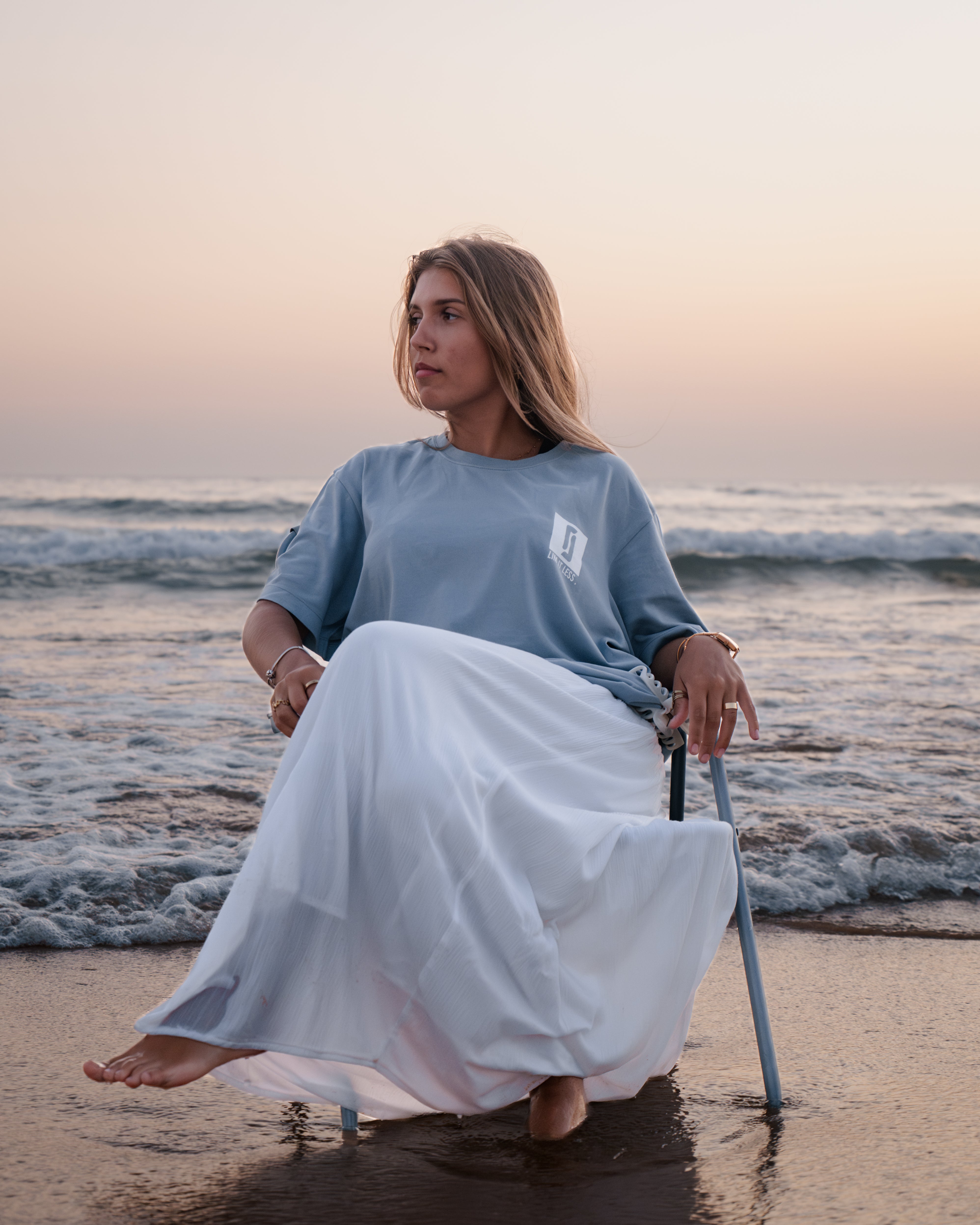 Modelo feminina com T-shirt Limitless Baía azul acinzentado em algodão premium, sentada em cadeira junto ao mar ao pôr-do-sol, coleção Modern Royalty.